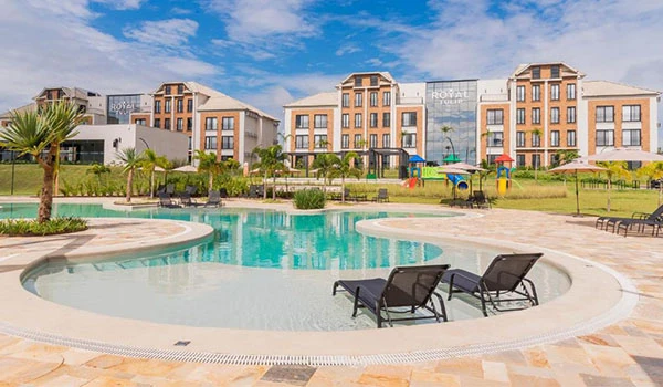 Imagem de uma piscina resort com espreguiçadeiras, jardim bem cuidado e edifícios modernos ao fundo, sob céu azul claro, ideal para relaxamento e lazer.