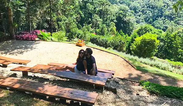 Casal sentado em banco de madeira ao ar livre em um parque com muita vegetação, árvores verdes ao fundo, ideal para fotos e momentos de lazer na natureza.