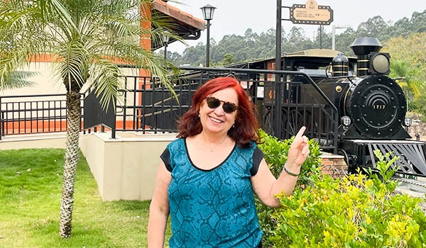 Deusa Rodrigues sorrindo e apontando para uma locomotiva a vapor ao fundo em um ambiente ao ar livre com árvores e plantas no Parque Três Pescadores em Aparecida.