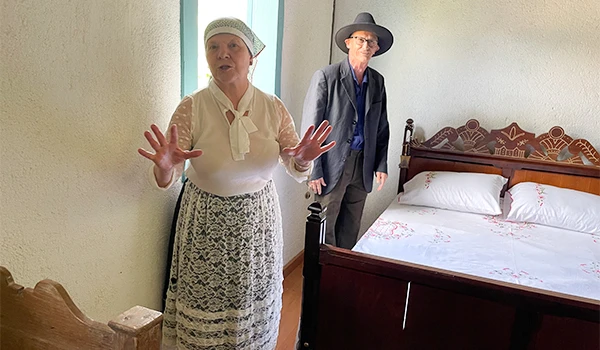 Imagem de uma cena em um quarto com uma mulher vestida de roupas tradicionais e um homem ao fundo. Ambos parecem apresentar algo, com a mulher gesticulando. Cena da interação na Casa Nostra na Rota das Montanhas Capixabas.