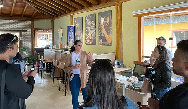 Imagem de um grupo em um ambiente acolhedor, onde uma mulher está apresentando uma ideia ou produto, em um evento de networking. O espaço é bem decorado e há interação entre os participantes.