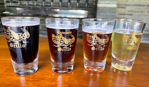 Imagem de 4 copos de cerveja Dus Grillo, apresentando diferentes estilos da bebida, em uma bancada de madeira.