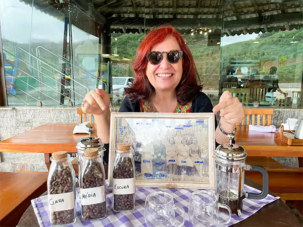 Deusa Rodrigues e usando óculos escuros em um café, com frascos de café ao seu redor e uma imagem emoldurada ao fundo. Cena de uma degustação na Rota das Montanhas Capixabas.