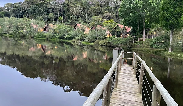 Ponte de madeira levando a casinhas em meio à natureza refletidas no lago calmo. Hotel Natureza Eco Lodge, na Rota das Montanhas Capixabas.