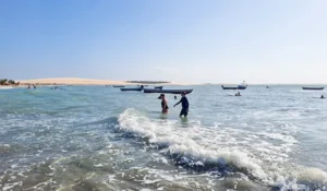 Imagem de uma praia com pessoas se divertindo nas ondas e barcos ao fundo. Um dia ensolarado destaca a beleza do mar e da areia.