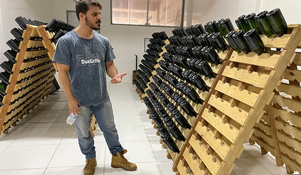 Homem em um ambiente de produção de vinhos, em frente a prateleiras de garrafas de vinho organizadas em madeira. A imagem ilustra o processo de vinificação.