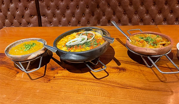 Imagem de três pratos de moqueca servidos em recipientes tradicionais sobre uma mesa de madeira. Os pratos oferecem uma variedade de cores e ingredientes.