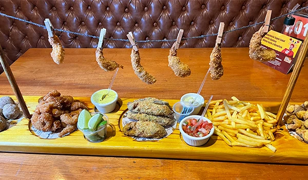 Prato de comida com diversas porções, incluindo camarão frito em um pequeno varal e abaixo batatas fritas, peixes e molhos, apresentados de forma atrativa,