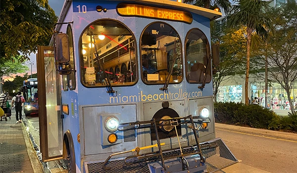 Trolebus Collins Express de Miami Beach em uma noite iluminada, mostrando o site miamibeachtrolley.com e uma atmosfera vibrante do local.