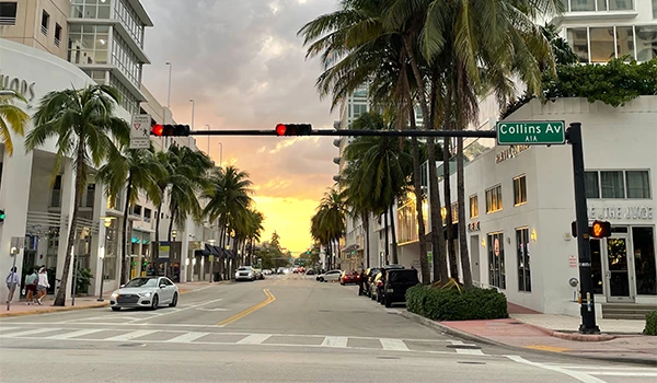 Imagem da Collins Avenue em Miami, com palmeiras e um pôr do sol no fundo. A rua apresenta um cenário urbano tranquilo e vibrante.