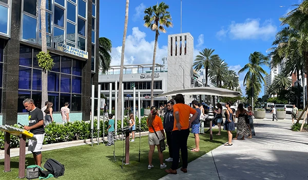 Imagem de um dia ensolarado com pessoas em um espaço público, cercadas por palmeiras e um edifício moderno ao fundo. Atrações de South Beach.