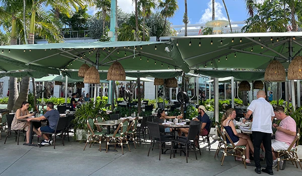 Ambiente de restaurante ao ar livre com mesas, cadeiras e um garçom atendendo clientes em um dia ensolarado. Plantas e decoração tropical ao fundo.