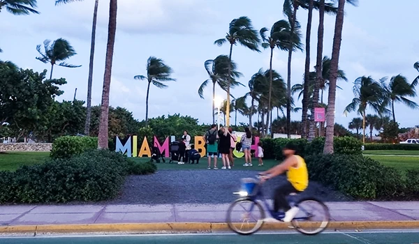 Pessoas posando em frente ao letreiro "Miami Beach" em um parque com palmeiras ao fundo, enquanto um ciclista passa. Uma das atrações de South Beach.