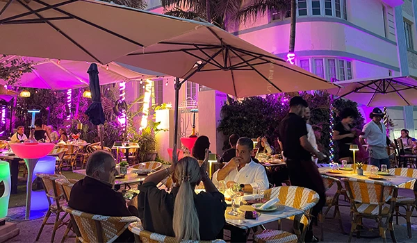 Imagem noturna de um restaurante iluminado com luzes coloridas e mesas cobertas por guarda-sóis. Atrações de South Beach.