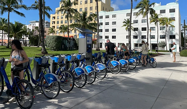 Imagem mostrando um ponto de aluguel de bicicletas em um ambiente urbano, com várias bicicletas azuis e pessoas utilizando o serviço. Uma das atrações de South Beach.