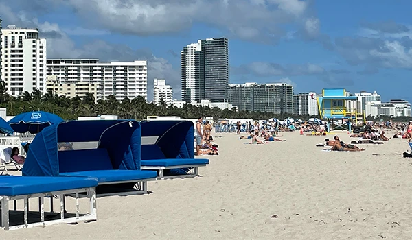 Praia de Miami com areia clara, quiosques azuis e prédios altos ao fundo. Um dia ensolarado cheio de pessoas aproveitando o mar e a areia.