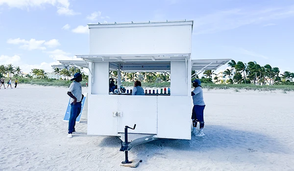 Quiosque de vendas na praia com vendedores e clientes, rodeado por palmeiras e areia, representando o lazer e o verão. Uma das atrações de South Beach.