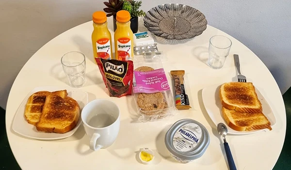 Café da manhã delicioso com torradas, bebidas e acompanhamentos em mesa branca. Itens incluem sucos, biscoitos, creme de queijo e café.