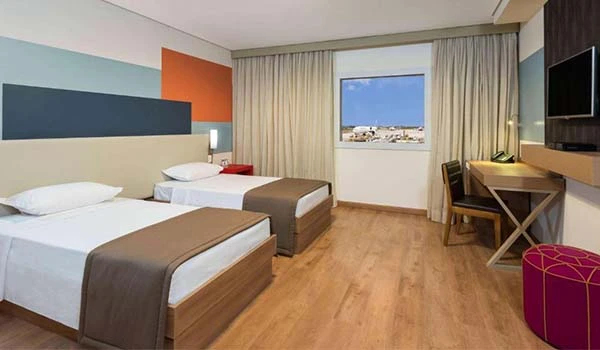 Quarto de hotel moderno com duas camas, decoração em cores suaves e vista para a cidade. Ideal para uma estadia confortável. Opção para utilizar também o estacionamento em Guarulhos Airport Park.