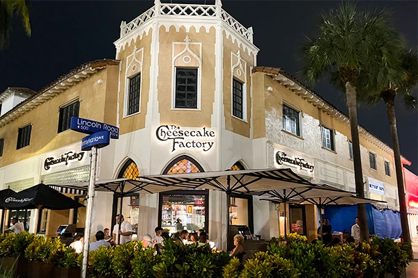 Imagem da Cheesecake Factory à noite, com mesas do lado de fora e uma bela arquitetura. O local é popular para jantar e sobremesas.