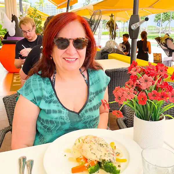 Deusa Rodrigues, usando óculos escuros, sorrindo em um restaurante ao ar livre. Ela está sentada à mesa com um prato delicioso e flores.