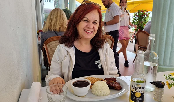 Deusa Rodrigues em um restaurante, com um prato de comida que inclui arroz, feijão e linguiça, além de uma cerveja Corona ao lado.