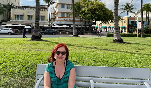 Deusa Rodrigues sentada em um banco no Lummus Park, com palmeiras e edifícios ao fundo. Atrações de South Beach.