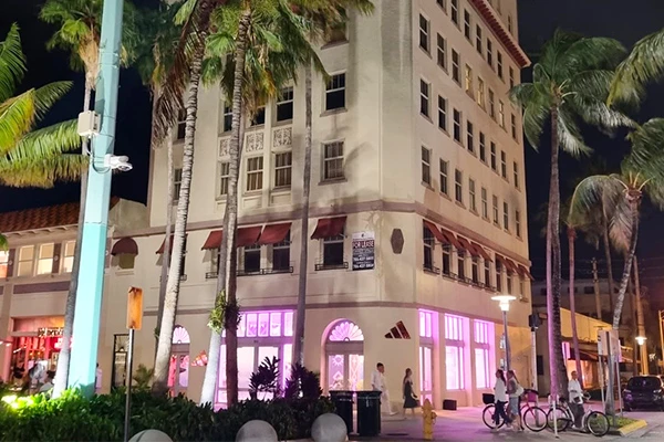 Imagem noturna de um edifício em Miami com iluminação rosa, cercado por palmeiras. O prédio exibe arquitetura clássica e um ambiente vibrante.