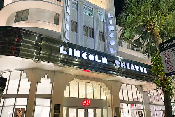 Uma imagem noturna do Lincoln Theatre, destacando sua arquitetura icônica e as letras iluminadas, hoje é sede de uma loja da HM em South Beach.