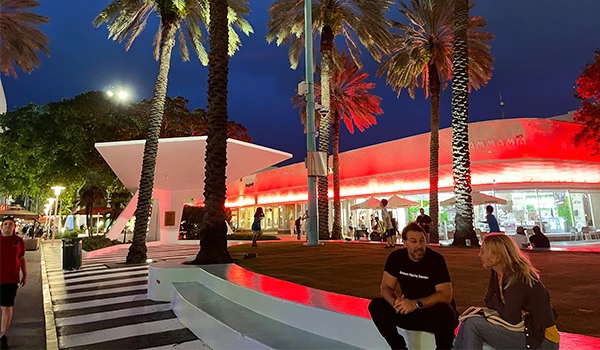 Imagem noturna de uma praça vibrante, com palmeiras iluminadas, pessoas socializando e um moderno edifício com fachada vermelha em destaque.