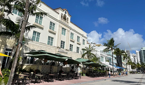 imagem da arquitetura de South Beach com mesas ao ar livre e palmeiras na calçada sob um céu azul. 