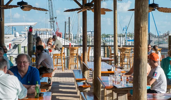 Ambiente descontraído em um bar à beira-mar com mesas e cadeiras de madeira. Clientes aproveitam a vista e a companhia em um dia ensolarado.