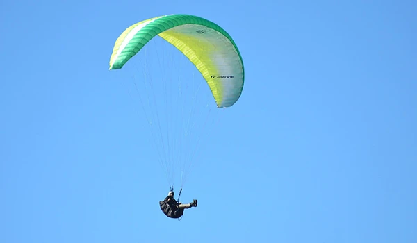 Um parapente colorido verde e amarelo flutua no céu azul enquanto um piloto desce suavemente, destacando a prática de parapente em locais ao ar livre.