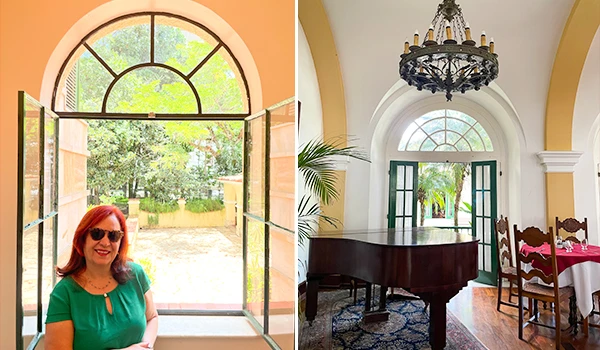 Deusa Rodrigues junto a uma janela aberta, com vista para um jardim verde. Ao fundo, um ambiente interno elegante com piano e decoração sofisticada.