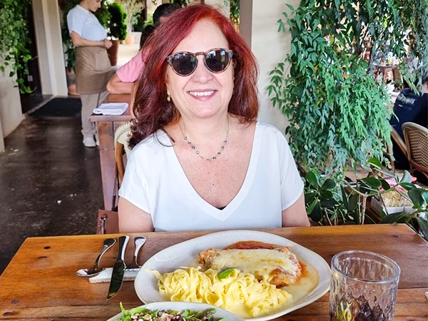 Deusa Rodrigues sorridente usando óculos escuros, sentada à mesa em um restaurante, com pratos de comida deliciosa à sua frente.