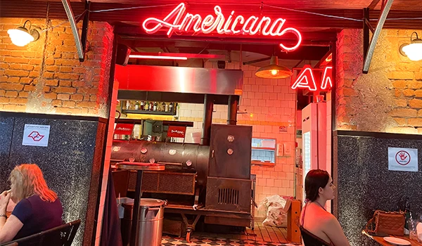 Vista da entrada do bar Americana, destacando a iluminação em néon e a cozinha ao fundo, ideal para quem busca um ambiente aconchegante e convidativo.