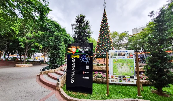 Praça de Serra Negra em São Paulo com árvores e decoração natalina, mostrando placa informativa sobre a cidade e um mapa, fica próximo da Fontana di Trevi.