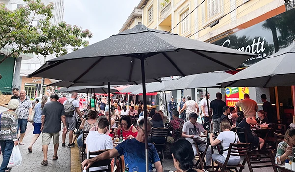 Imagem de um movimentado restaurante ao ar livre com mesas sob guarda-sóis, pessoas socializando e aproveitando o ambiente. Animação e descontração em véspera de feriado.