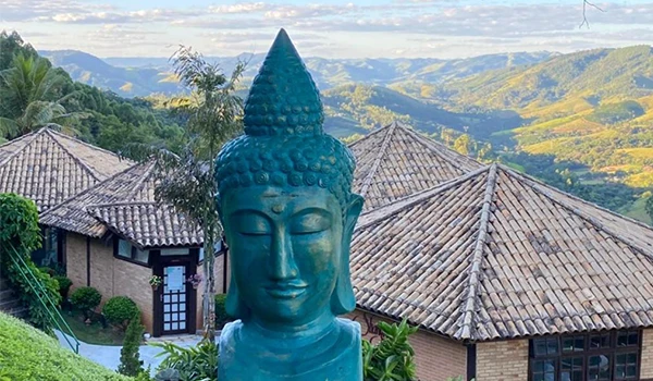 Foto da Pousada Shagri-la com escultura de Buda em meio à natureza, com vista para montanhas e casas com telhado de cerâmica. Um ambiente tranquilo para meditação e relaxamento.