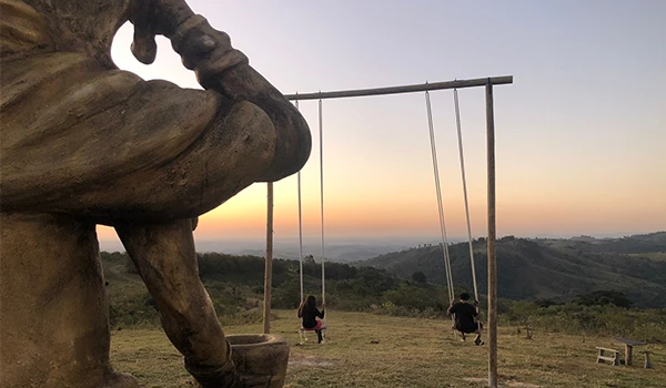 Escultura de um homem pensativo em primeiro plano com crianças brincando em balanços ao pôr do sol, em um cenário natural de montanhas.