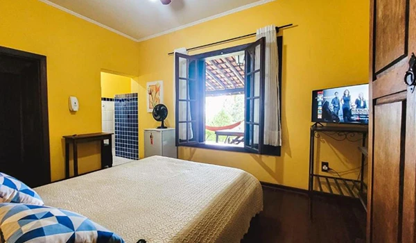 Quarto aconchegante com paredes amarelas, cama confortável, ventilador, janela ampla e TV, ideal para relaxamento e descanso em um ambiente tranquilo.