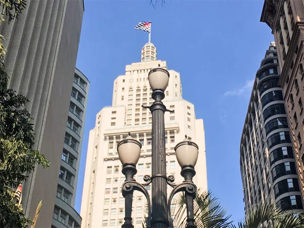 Imagem do Edifício Altino Arantes, em São Paulo, com a bandeira do Brasil no topo e postes de luz em primeiro plano.