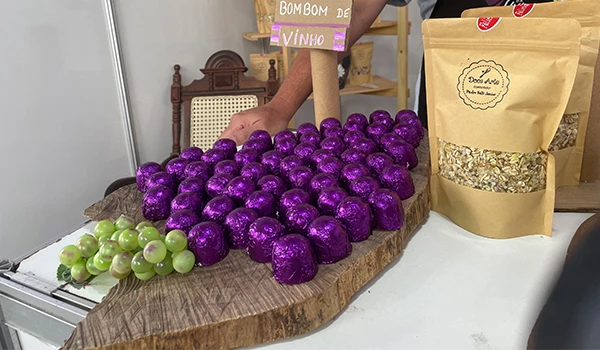 Exibição de bombons de vinho em uma mesa de madeira, com bombons embrulhados em papel roxo, acompanhados por uvas verdes e pacotes de produtos naturais.