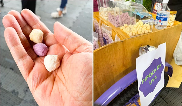 Pipoca de uva em um estande de rua, com uma mão segurando três pedaços coloridos. O letreiro ao fundo indica o sabor da pipoca.