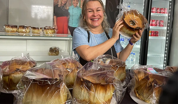 Mulher sorridente segurando um bolo decorado em uma loja com outros bolos ao fundo. A imagem destaca produtos de confeitaria e o sorriso da proprietária.