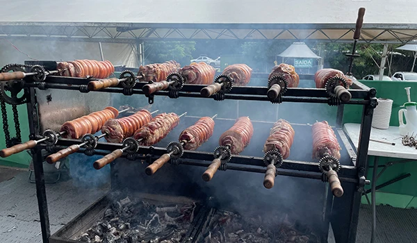 Espetinhos de carne assados em fogo de chão com fumaça, tradição de churrasco brasileiro, em um evento ao ar livre.