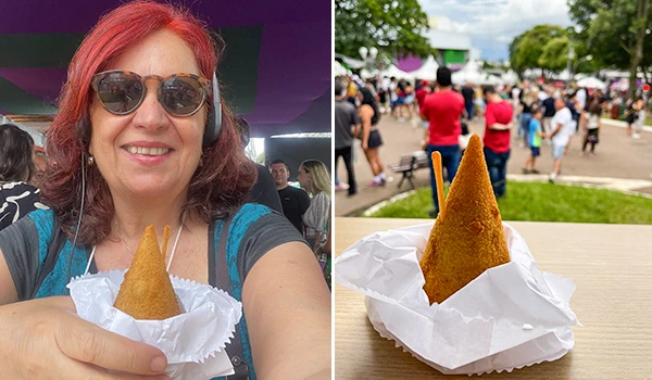 Deusa Rodrigues degustando uma coxinha de queijo em um evento lotado, transmitindo alegria e descontração.