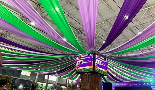 Imagem de um evento decorado com faixas verdes e roxas penduradas no teto, criando um ambiente festivo e colorido. Painéis informativos também estão visíveis, na típica Festa da Uva de Jundiaí.