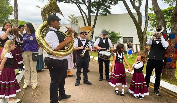 Banda de música tocando em evento ao ar livre, com músicos e crianças vestindo trajes tradicionais. Atmosfera festiva e vibrante.