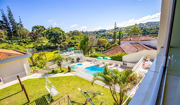 Vista panorâmica de uma área de lazer com piscina, jardim e espreguiçadeiras em um dia ensolarado.